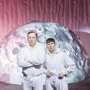 Dwóch mężczyzn w białym dresie ze strapionymi minami stoi przed rekwizytem dużego księżyca. Ściana w tle imituje skały.