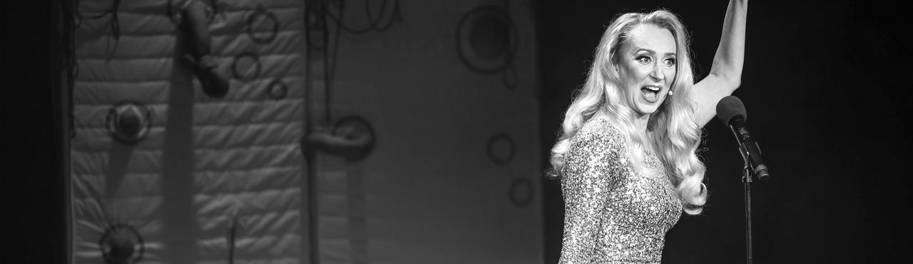 Kobieta w blond włosach i cekinowej sukience śpiewa do mikrofonu. Lewą rękę trzyma podniesioną do góry.