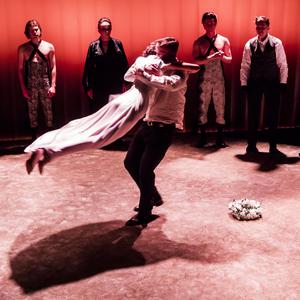 Mężczyzna obraca w tańcu kobietę w sukni ślubnej, przyglądają im się ludzie w eleganckich kostiumach. Na podłodze leży wianek.