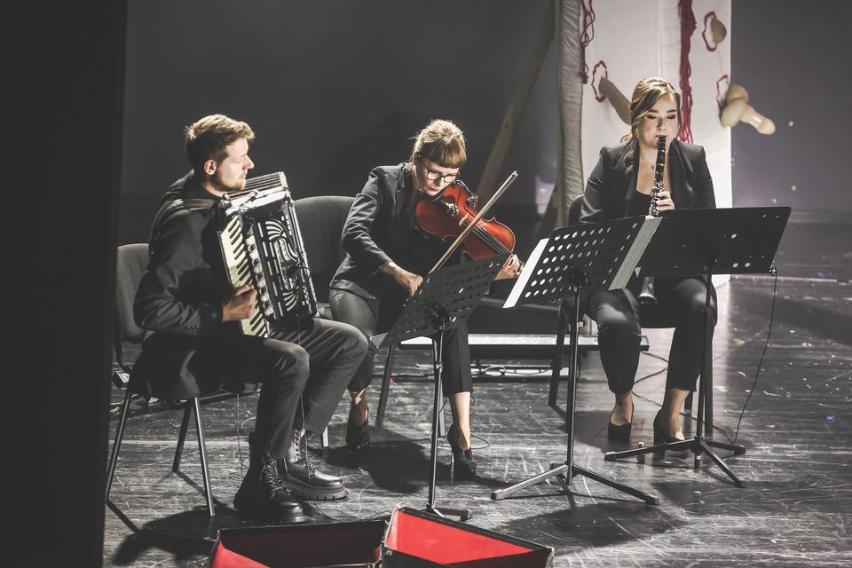 Troje muzyków siedzi na krzesłach przy pulpitach. Mężczyzna z lewej trzyma akordeon, dwie kobiety mają altówkę oraz klarnet.